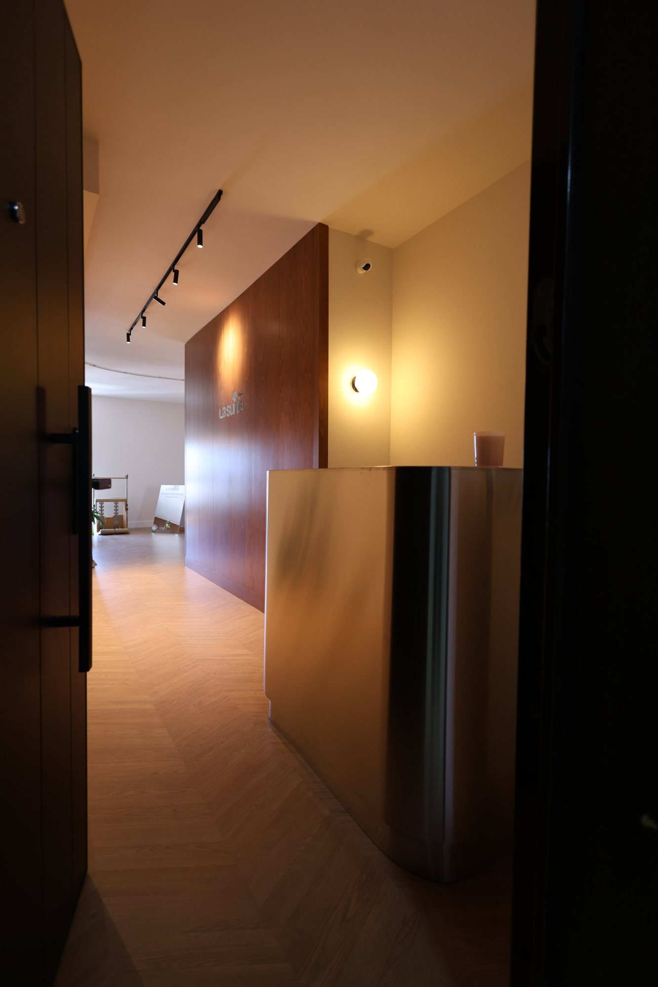 View through the entrance of La Suite toward the folded stainless steel reception desk, curved at its corner beside an oak partition