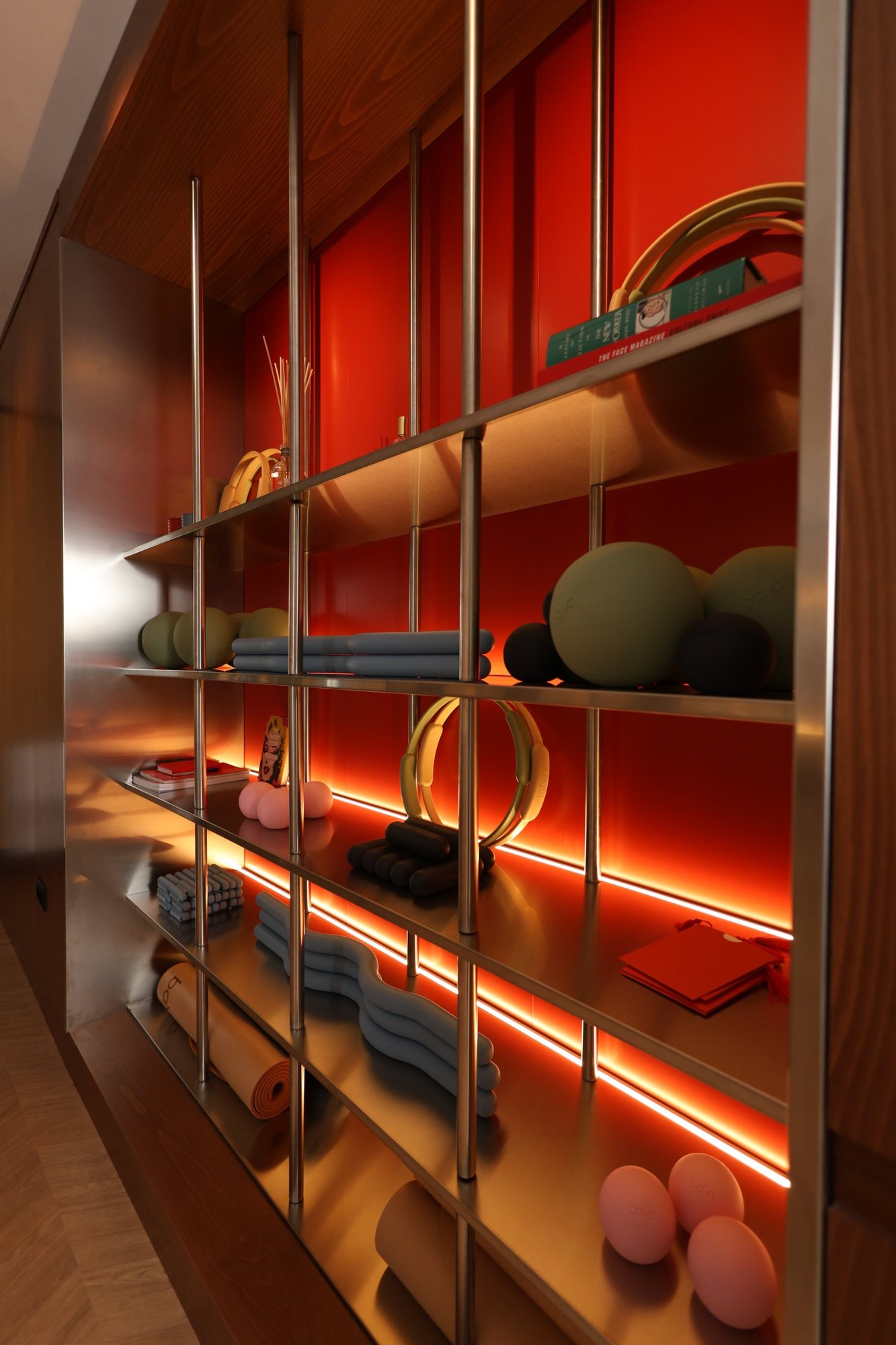 Angled close view of polished stainless steel uprights and illuminated trays holding pilates balls, rings and mats against an orange backing at La Suite