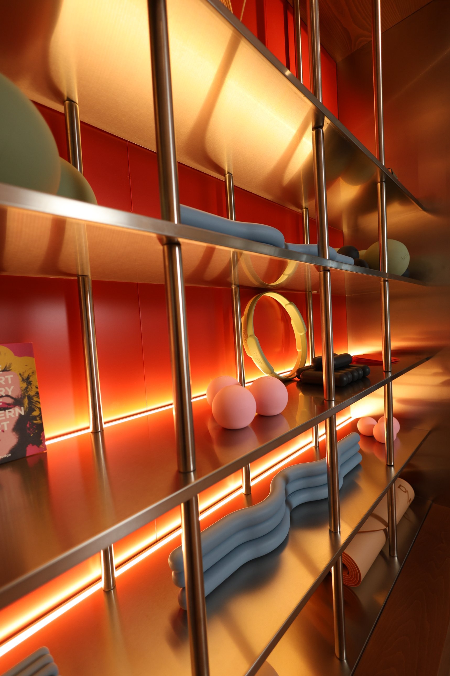 Close-up of the brushed stainless steel shelves and polished uprights, with pilates balls, rings and wave rollers, lit from below against an orange wall at La Suite